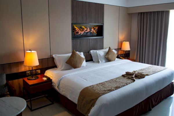 A cozy hotel room with a large bed, two side tables with lamps, a wall-mounted fireplace, and a window with curtains.
