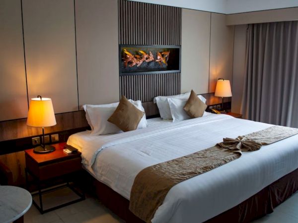 A cozy hotel room with a large bed, two side tables with lamps, a wall-mounted fireplace, and a window with curtains.