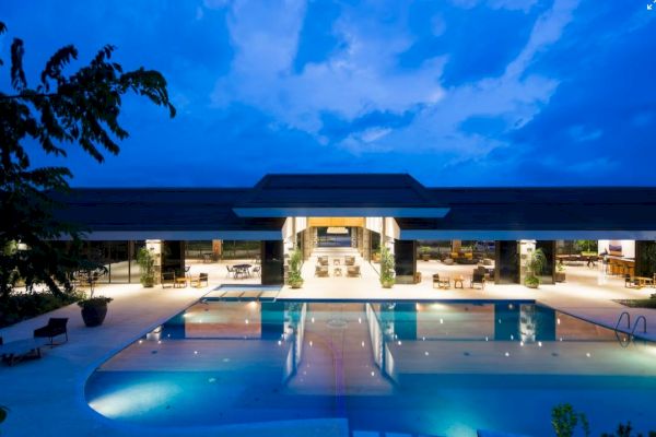 The image shows a modern, luxurious building with a large swimming pool at night, beautifully lit under a deep blue sky with scattered clouds.