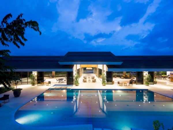 The image shows a modern, luxurious building with a large swimming pool at night, beautifully lit under a deep blue sky with scattered clouds.