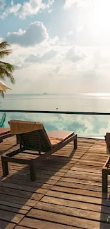 Poolside relaxation area with lounge chairs, umbrellas, and a stunning view of a calm water body surrounded by palm trees at sunrise or sunset.