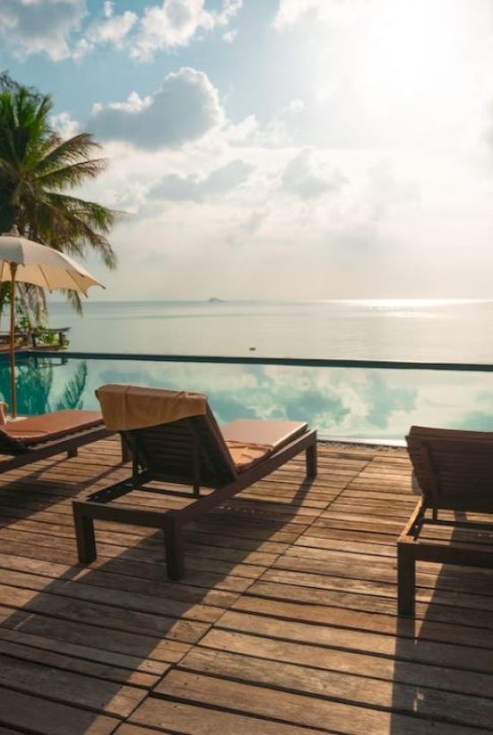 A serene poolside scene with lounge chairs and umbrellas, overlooking a calm ocean, under a partly cloudy sky and surrounded by lush trees.