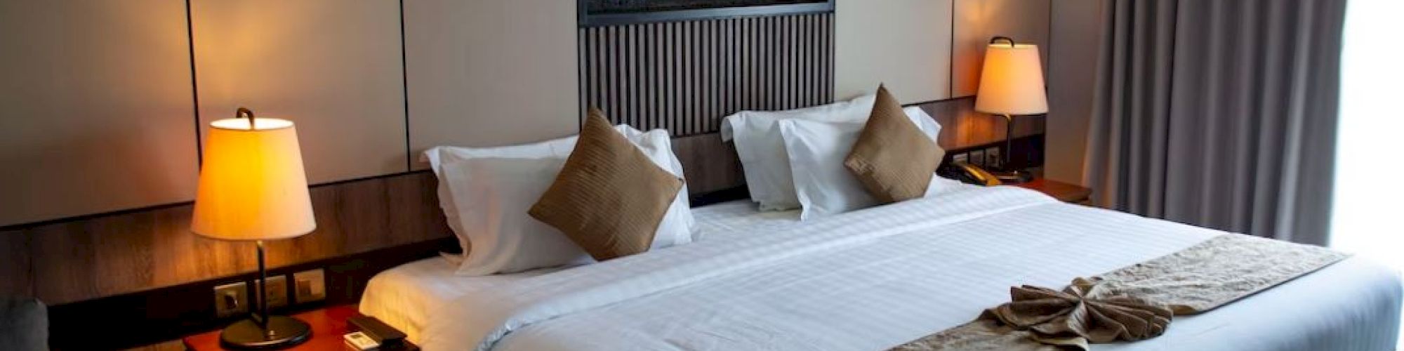 This image shows a well-lit hotel room featuring a large bed with brown throw pillows, bedside lamps, a wall-mounted fireplace, and a large window with curtains.