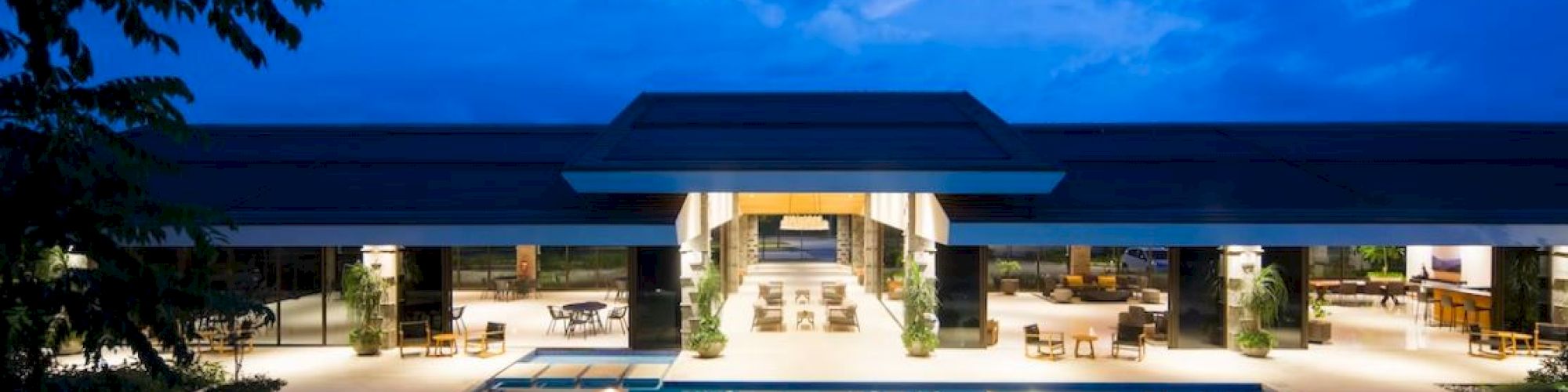 A luxurious outdoor pool area at dusk, surrounded by a modern building with open seating and glowing lights, set against a blue sky.