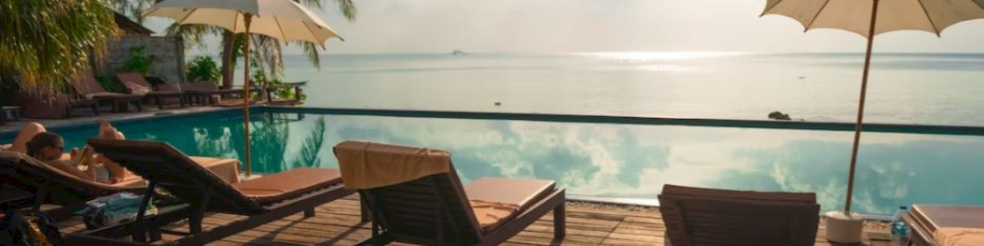 A serene tropical poolside scene with lounge chairs, umbrellas, and palm trees, overlooking a calm coastline under a bright, partially cloudy sky.