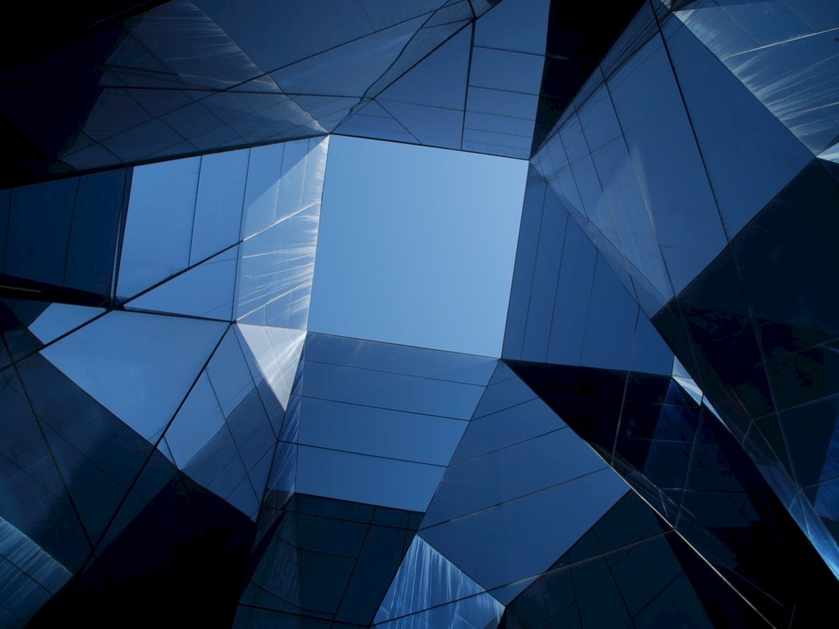 A geometric pattern of reflective glass surfaces creating a kaleidoscopic effect, with a clear blue sky seen through the center.