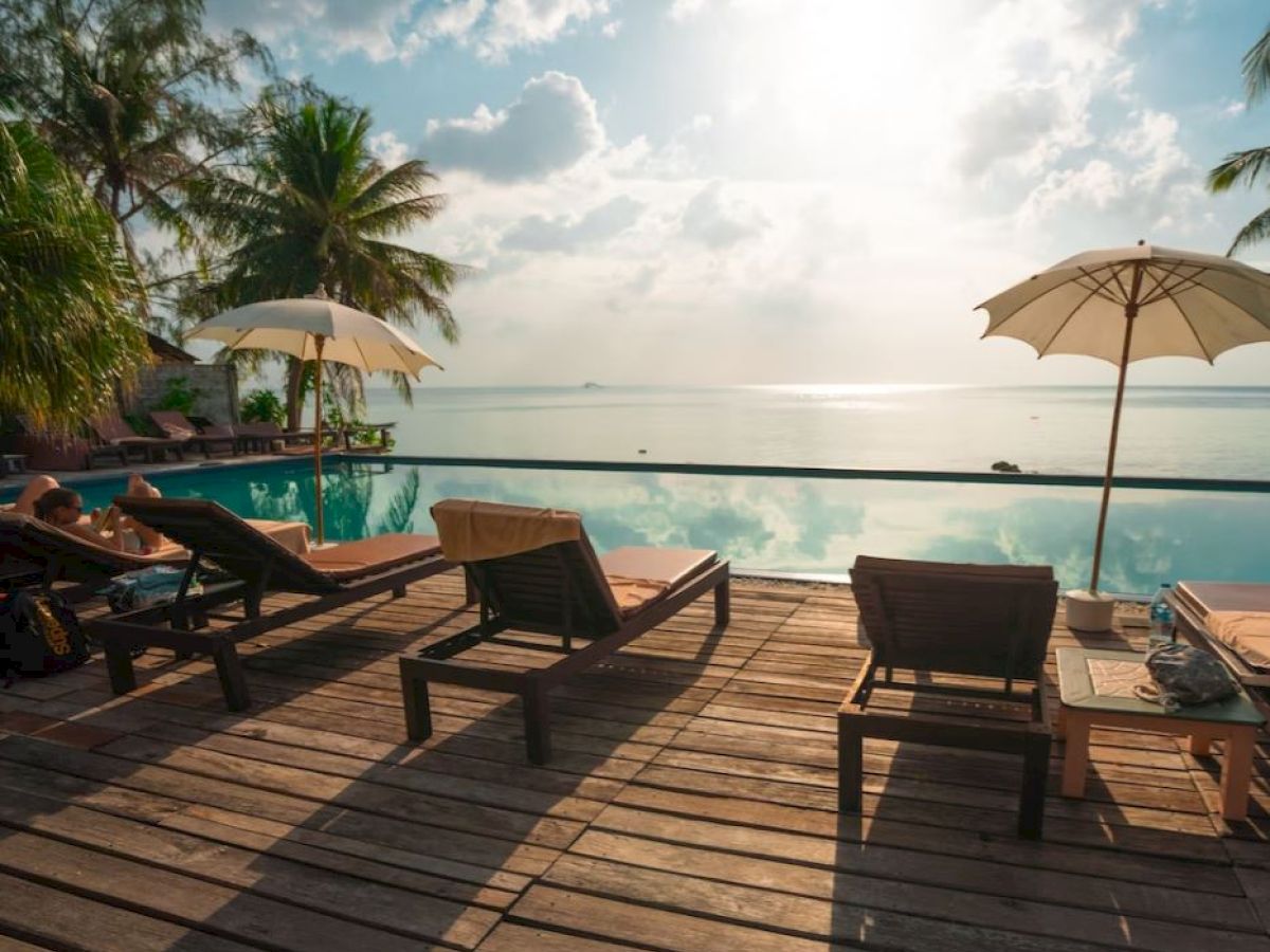 A serene poolside scene with lounge chairs and umbrellas under a bright sky, overlook the calm water, surrounded by lush palm trees.