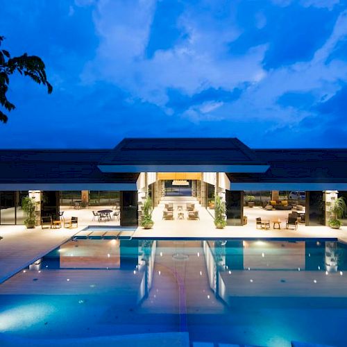 The image features a luxurious building with an expansive, illuminated outdoor swimming pool during dusk or early evening, framed by a vibrant sky.