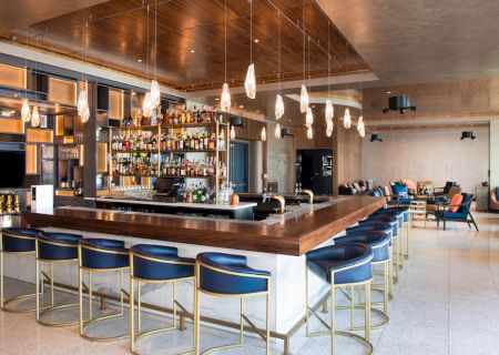 The image shows a modern bar with a wooden counter, bar stools, a variety of liquor bottles, pendant lighting, and seating areas in the background.