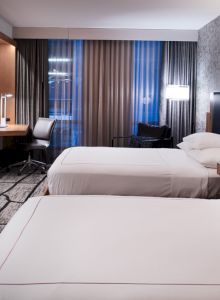 This image shows a modern hotel room with two neatly made beds, a desk, a flat-screen TV, and contemporary decor and lighting.