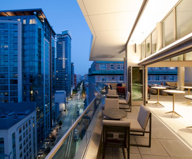 A modern cityscape at dusk from a high-rise balcony with seating and tables, overlooking a street flanked by tall buildings and illuminated.