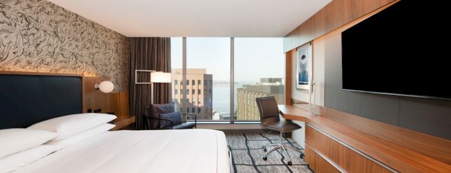 A modern hotel room with a large bed, a wall-mounted TV, a desk, a chair, and large windows offering a city and water view.
