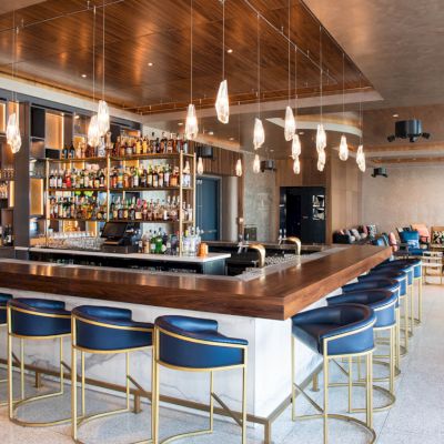 The image shows a modern bar with a wooden counter, blue chairs, pendant lights, and a well-stocked back bar. The seating area is visible in the background.