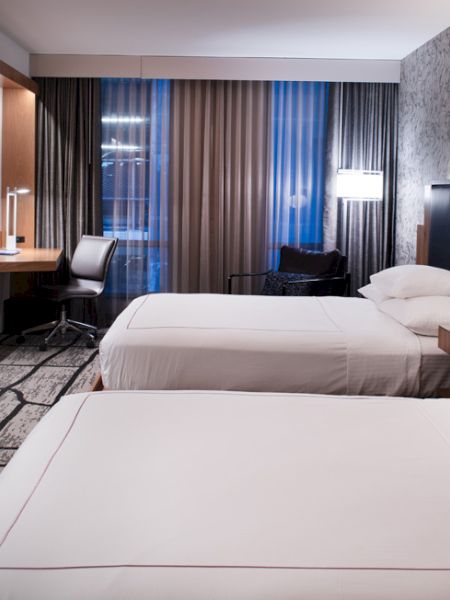 The image shows a modern hotel room with two beds, a desk, a flat-screen TV, and contemporary decor. The windows are covered with blinds.