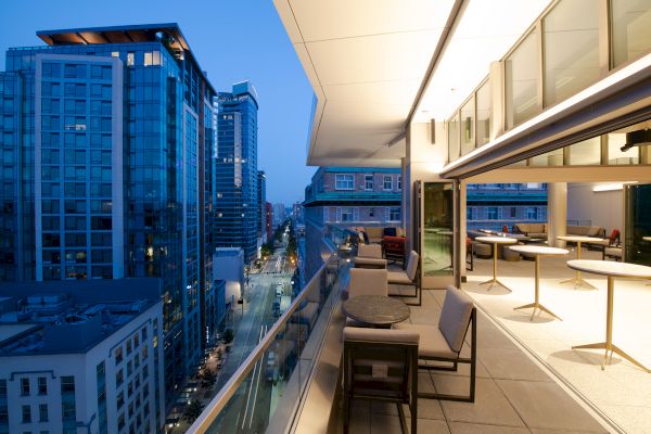 A modern rooftop terrace with tables and chairs, overlooking a city street flanked by tall buildings at dusk.