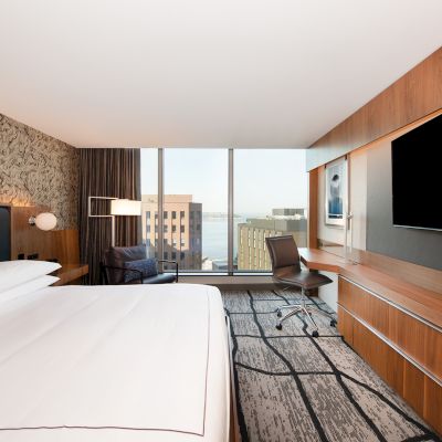 A modern hotel room with a large bed, flat-screen TV, desk, chair, and large windows overlooking buildings and water.