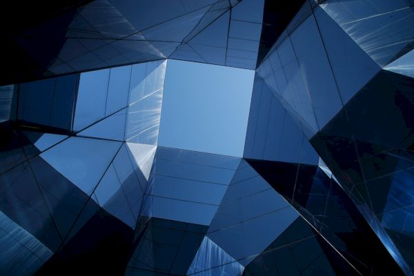 This image shows abstract architecture with reflective glass panels forming geometric shapes, looking up to a blue sky in the center of the composition.