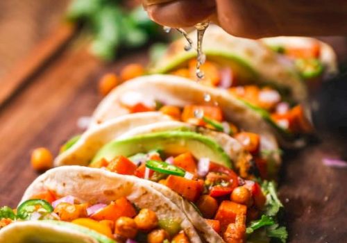 The image shows a hand squeezing lime juice over a row of deliciously prepared tacos filled with vegetables and chickpeas on a wooden surface.