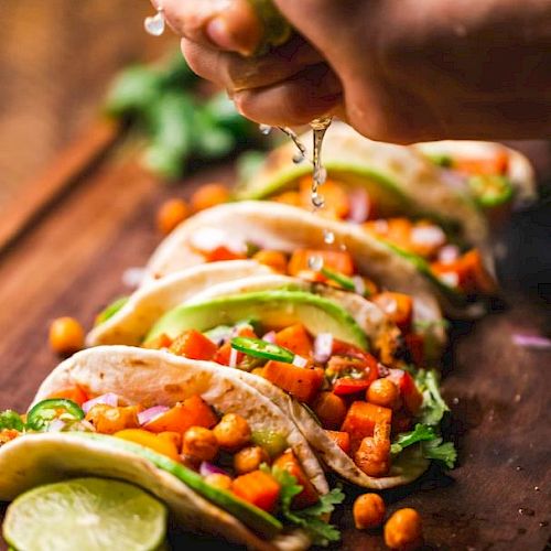 The image shows a hand squeezing lime juice over a row of deliciously prepared tacos filled with vegetables and chickpeas on a wooden surface.