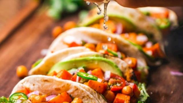 The image shows a hand squeezing lime juice over a row of deliciously prepared tacos filled with vegetables and chickpeas on a wooden surface.