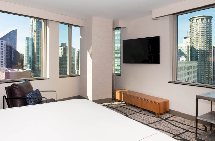 A modern hotel room with large windows showcasing a cityscape, a wall-mounted TV, a chair, and a bed.