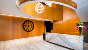A modern lobby with wood paneling, a large clock, a marble front desk, a decorative light fixture, and a vase of flowers.