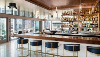 A stylish bar with a wooden countertop, gold-trimmed barstools, and a large selection of bottles behind the counter, with large windows and city views.