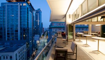 The image shows a modern cityscape from a high balcony with outdoor seating and tables, adjacent to a spacious indoor area.