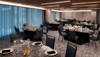 An elegant banquet hall with round tables set for an event, featuring black chairs, white floral centerpieces, and blue curtains in the background.