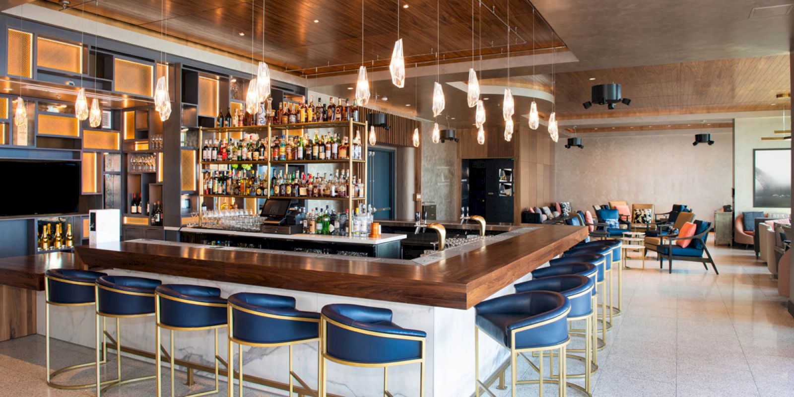 The image shows a modern bar with blue seats, a wooden countertop, hanging lights, and shelves stocked with various bottles. The background has lounge seating.