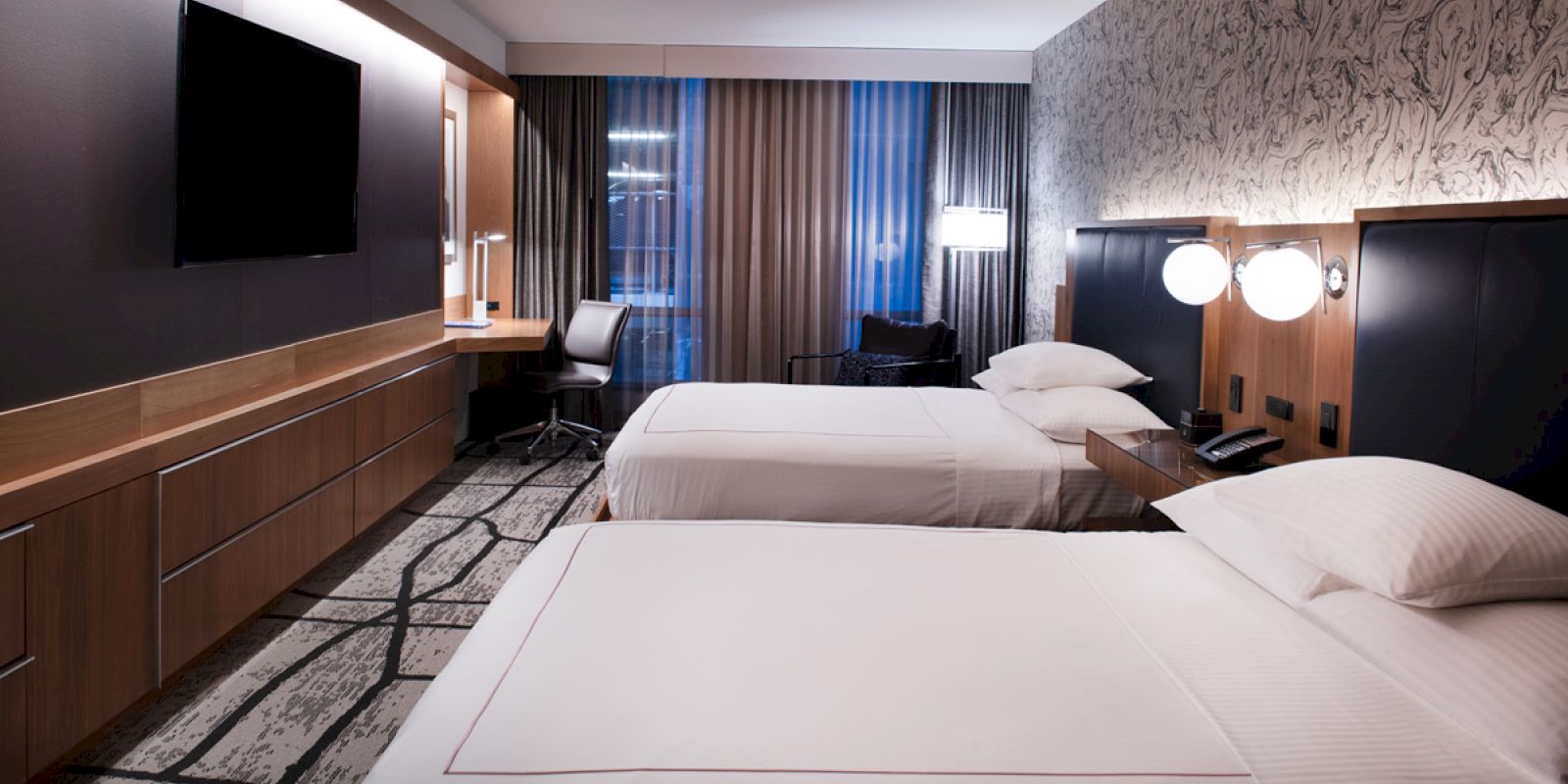 This image shows a modern hotel room with two large beds, a TV, a desk and chair, and contemporary decor with patterned walls and floor.