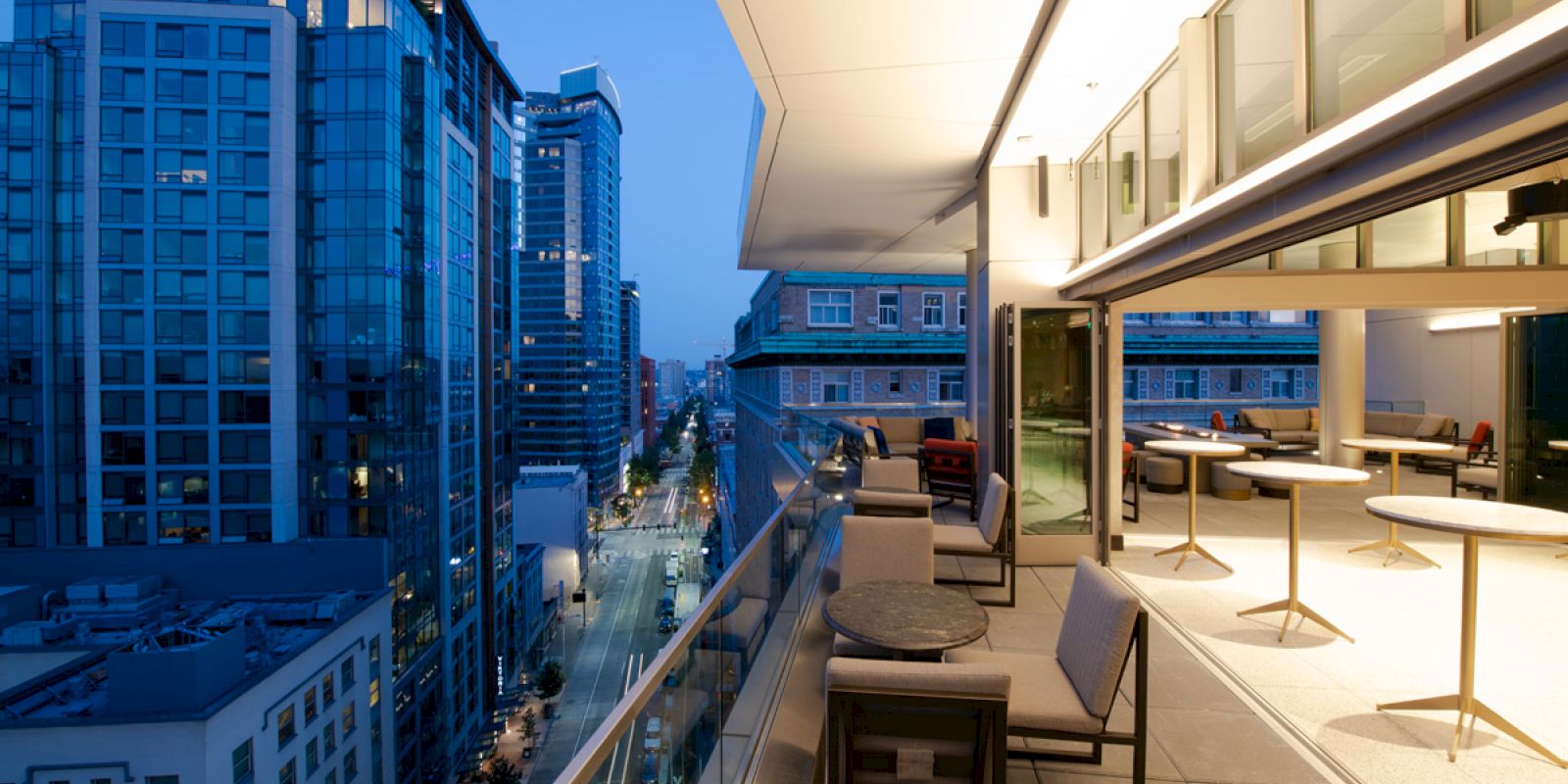 A modern urban balcony at dusk overlooking a city street, furnished with tables and chairs, adjacent to a building with large glass windows.