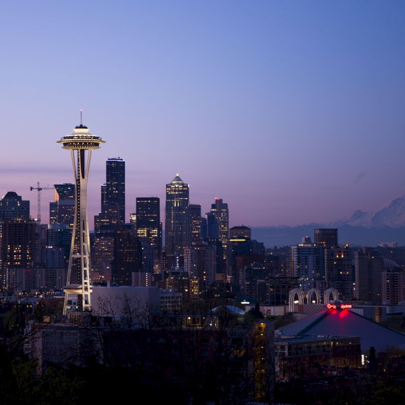 A cityscape featuring the Seattle skyline at dusk, with the Space Needle prominently visible and Mount Rainier in the background.