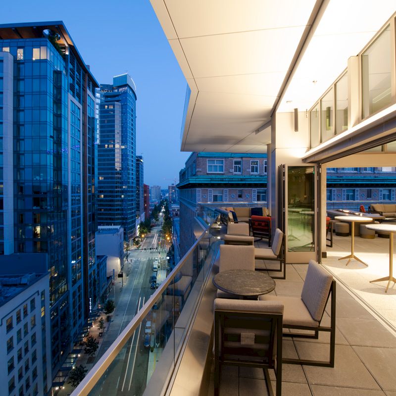 A modern rooftop terrace with tables and chairs overlooking a cityscape filled with tall buildings during twilight.