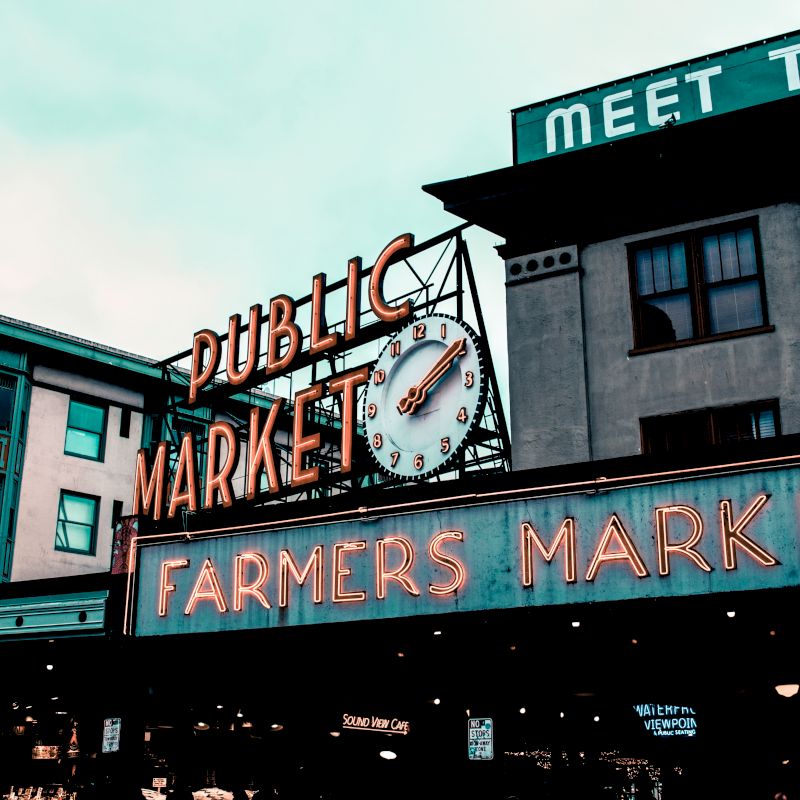 An outdoor neon sign reads "Public Market" and "Farmers Market" with a clock above it, located on a building in a marketplace.