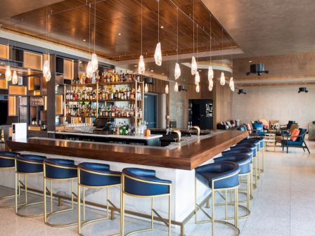 The image shows a modern bar area with a central counter, blue high chairs, hanging lights, and shelves stocked with bottles. There are seating areas too.