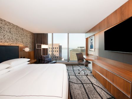 A modern hotel room with a large bed, a desk with a chair, a wall-mounted TV, and a large window offering a city view.