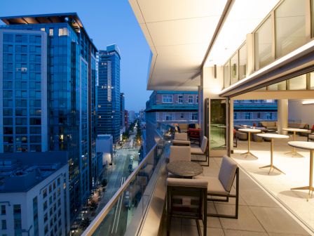 A modern, open-air balcony at dusk in a cityscape with outdoor seating, glass railings, and high-rise buildings visible in the background.