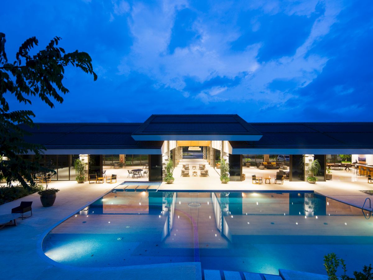 An illuminated building with a large, luxurious swimming pool at twilight, featuring open seating areas and a modern architectural design.