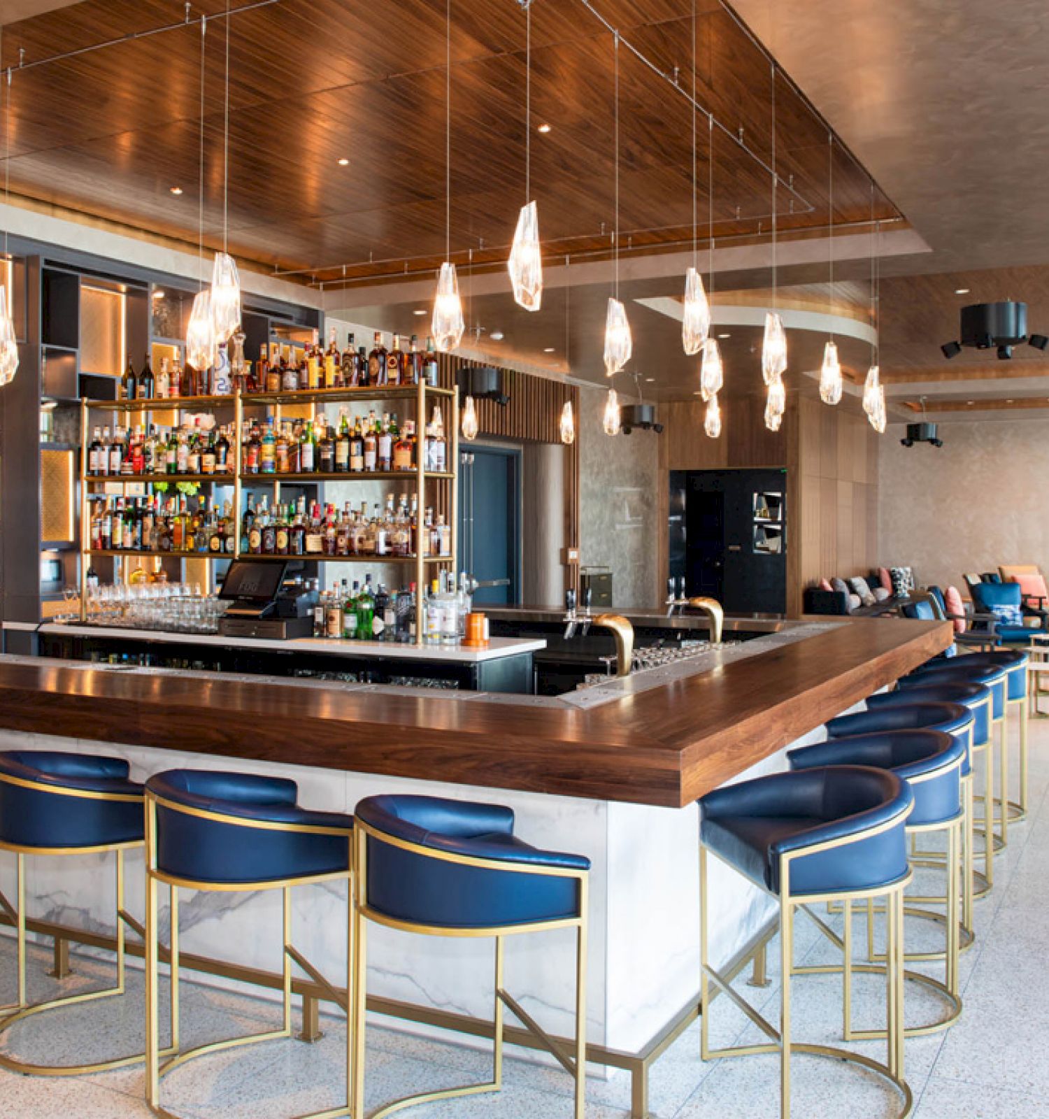 A modern bar with blue stools, a well-stocked liquor shelf, hanging lights, and a lounge area with seating in the background.