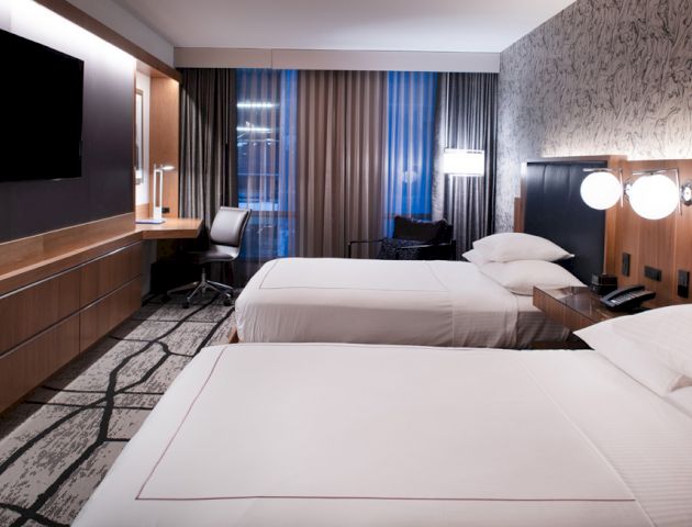 The image shows a modern hotel room with two double beds, a wall-mounted TV, a workstation with a chair, and stylish decor including a patterned accent wall.