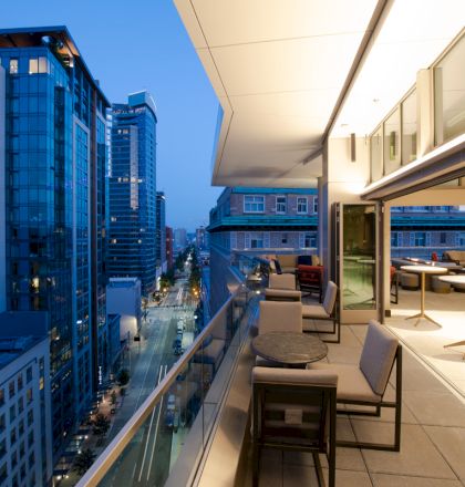A modern urban balcony overlooks a cityscape with tall buildings and lit streets at dusk. The balcony is furnished with tables and chairs.