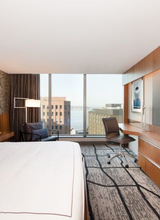 Modern hotel room with a large bed, flat-screen TV, desk, and chair. The window offers a cityscape view with some buildings in the background.