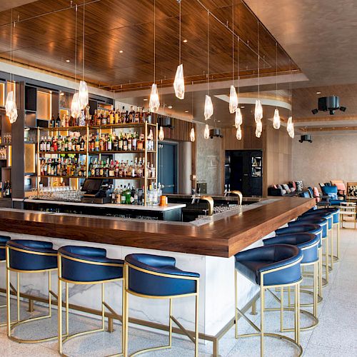 The image shows a modern bar with blue bar stools, a well-stocked bar, pendant lights, and a seating area with tables and chairs in the background.