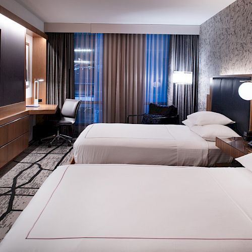 A modern hotel room with two neatly made beds, a flat-screen TV, a desk with a chair, and contemporary decor illuminated by soft lighting.
