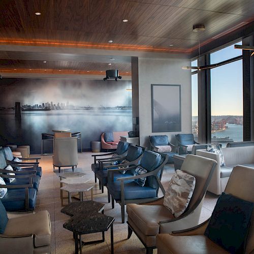A modern lounge area with comfortable seating, large windows offering a scenic view of the waterfront and a cityscape artwork on the wall.