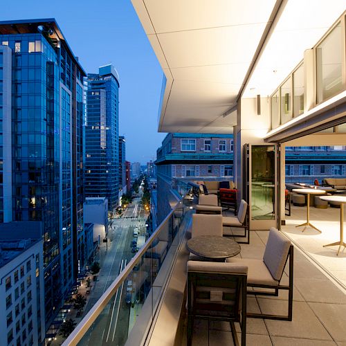 A modern balcony with seating areas overlooks a city street lined with tall buildings at twilight. The interior has tables and chairs.
