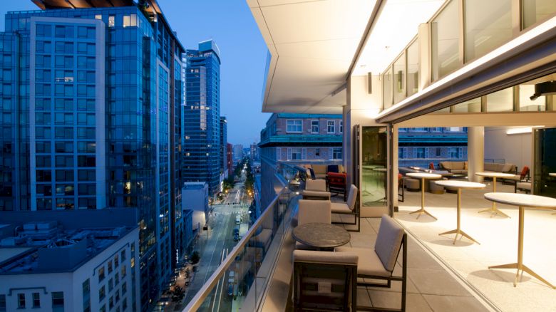 An outdoor balcony at dusk with modern furniture overlooks a cityscape of tall buildings and lit streets, providing a scenic urban view.