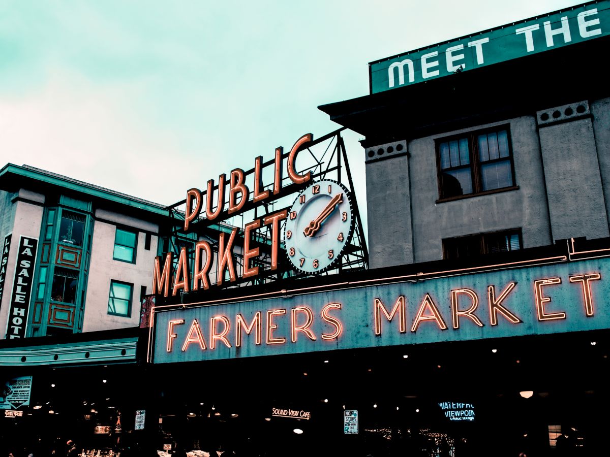 The image shows the entrance of a public farmers market with a vintage neon sign displaying "PUBLIC MARKET" and "FARMERS MARKET" below a clock.