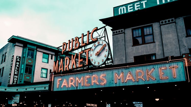 The image shows the entrance of a public farmers market with a vintage neon sign displaying "PUBLIC MARKET" and "FARMERS MARKET" below a clock.